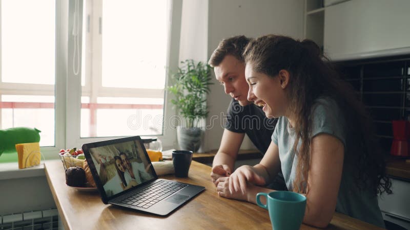 Die jungen netten kaukasischen Paare, die bei Tisch vor dem Laptop skyping ist mit ihrem glücklichen Positiv sitzen, bräunten Fre