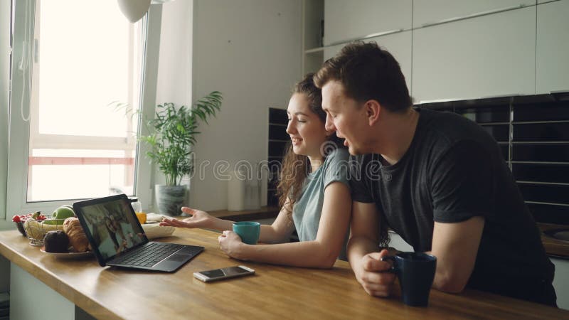 Die jungen netten kaukasischen Paare, die bei Tisch vor dem Laptop skyping ist mit ihrem glücklichen Positiv sitzen, bräunten Fre