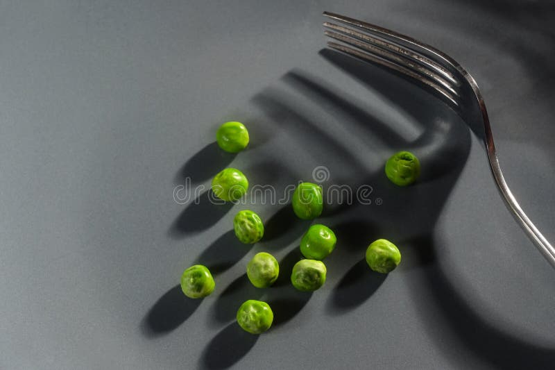 Hard diet with a few green peas and a fork with shadows on a gray background with copy space, modern abstract concept for resolution to eat vegetarian and slim down, copy space, selected focus. Hard diet with a few green peas and a fork with shadows on a gray background with copy space, modern abstract concept for resolution to eat vegetarian and slim down, copy space, selected focus