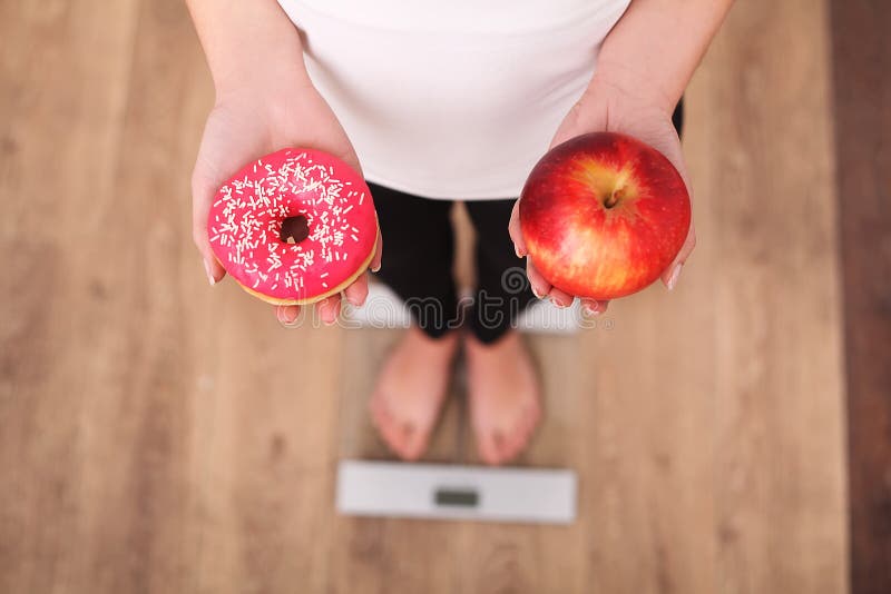 Dieta Peso corporal de medição da mulher na escala de peso que guarda a filhós e a maçã Os doces são comida lixo insalubre Fazend