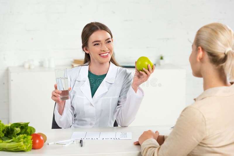 Dietititer i vit päls som håller äpple och glas vatten och patient på bordet royaltyfria foton