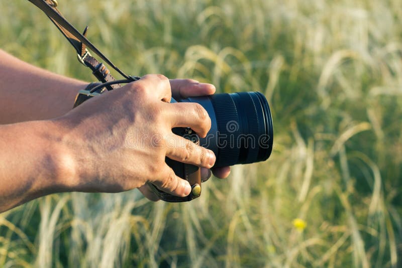 The digital DSLR camera in a hand. Male hands with camera take photo of green grass stock photo The digital DSLR camera in a hand. Male hands with camera take photo of green grass stock photo