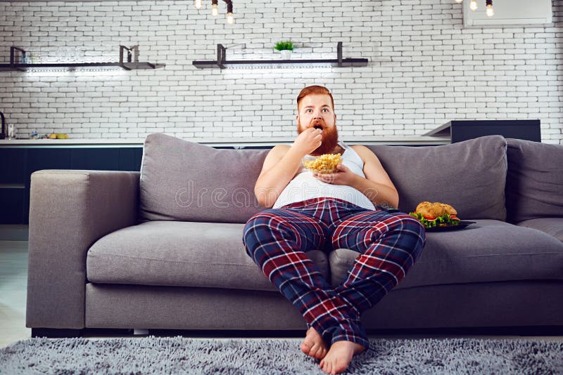 Dikke grappige mens die in pyjama's een hamburgerzitting op de laag eten royalty-vrije stock afbeelding Dikke grappige mens die in pyjama's een hamburgerzitting op de laag eten royalty-vrije stock afbeelding