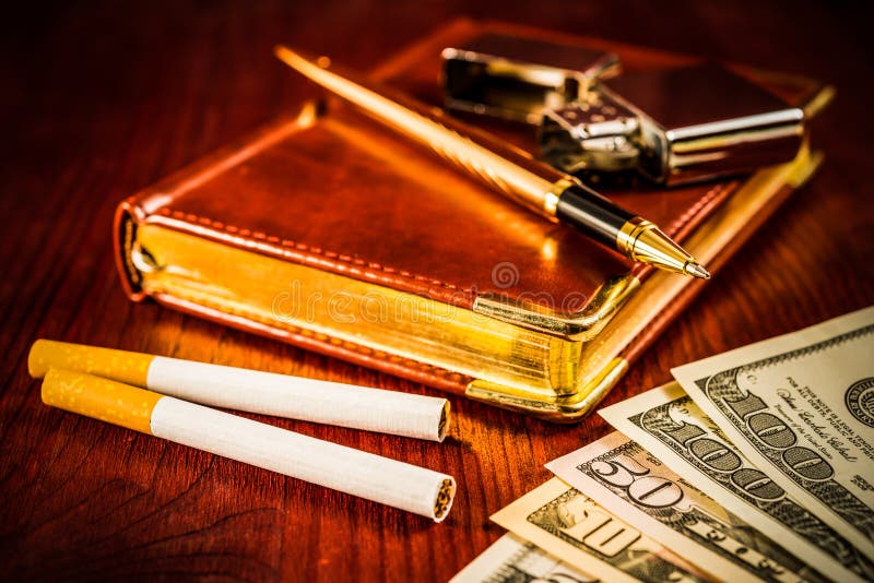 Money with a leather diary and cigarettes with golden pen on a mahogany table. Focus on the cigarettes. Money with a leather diary and cigarettes with golden pen on a mahogany table. Focus on the cigarettes