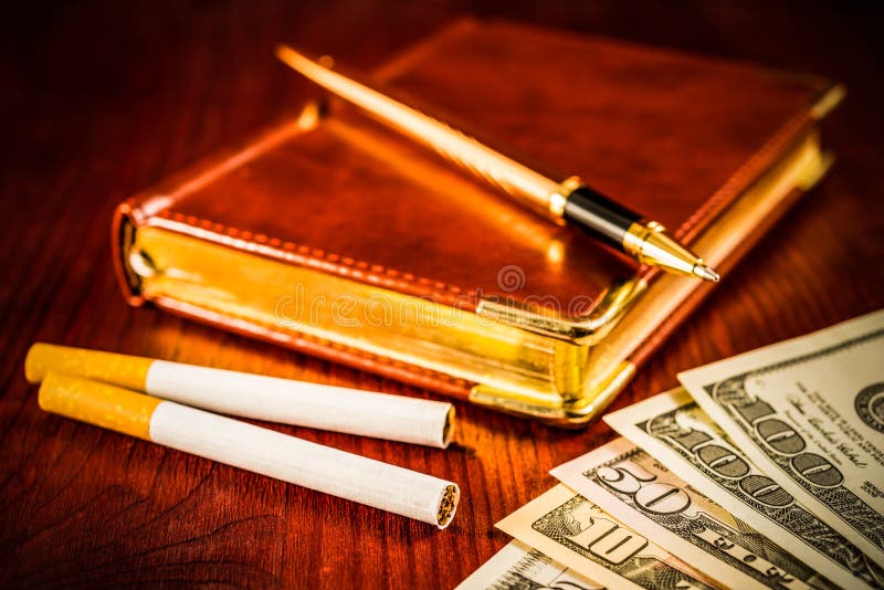Money with a leather diary and cigarettes with golden pen on a mahogany table. Focus on the cigarettes, shallow deep of field. Money with a leather diary and cigarettes with golden pen on a mahogany table. Focus on the cigarettes, shallow deep of field