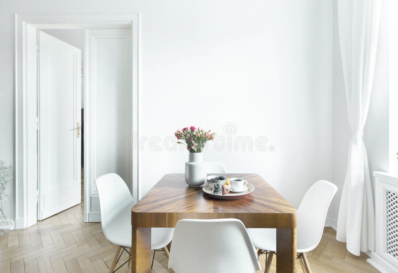 Dining table with fresh flowers and breakfast tray with coffee cup and fruits in real photo of white room interior with empty wall