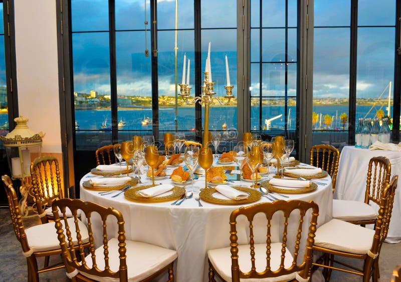 Wedding, party or business event tables decoration in white and golden tones. Silvery candelabrums and cutlery. Beautiful wide windows with urban bay view. Wedding, party or business event tables decoration in white and golden tones. Silvery candelabrums and cutlery. Beautiful wide windows with urban bay view.