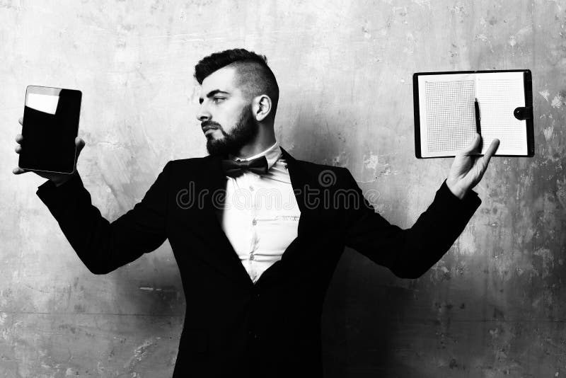 Business director with beard and calm face wearing stylish suit holds tablet and blank open organizer on old beige wall background. Concept of old fashioned and modern planning styles and management. Business director with beard and calm face wearing stylish suit holds tablet and blank open organizer on old beige wall background. Concept of old fashioned and modern planning styles and management