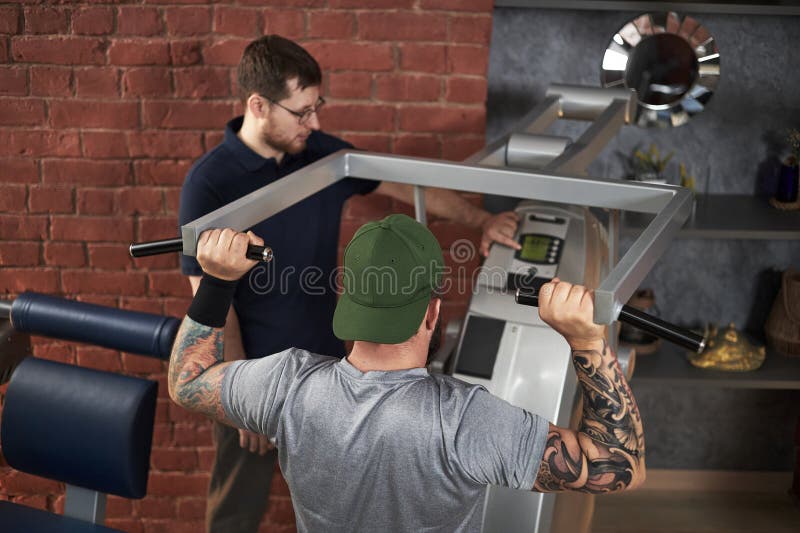 Male fitness instructor explaining proper technique for machine exercise to men in green cap in gym setting with brick wall background. Exercise technique stock images, royalty-free photos and pictures