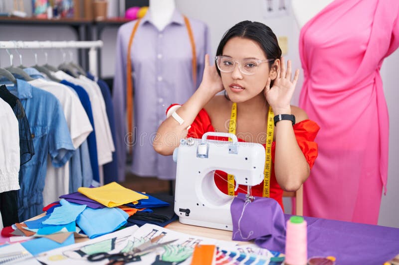 Diseñadora de costurera hispana que usa una máquina de coser para tratar de escuchar ambos gestos de oídos curiosos por los chisme fotos de archivo