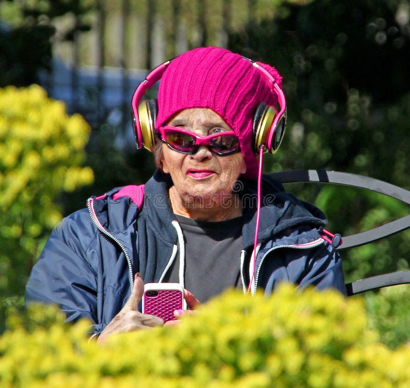 Fotografie mladé v srdci babička převlečený v růžový pletený klobouk a na sobě jogging tepláky a sluneční brýle, poslouchání stáhnout itunes na mobilní zařízení někde v Kent park! 