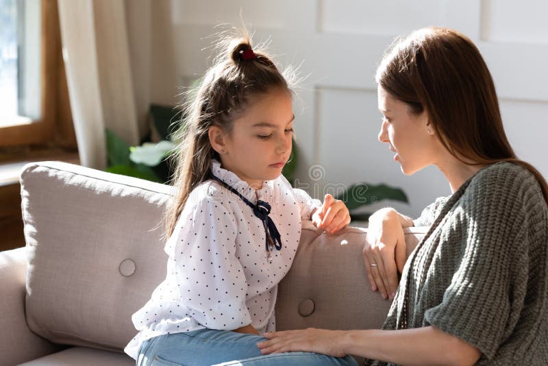 Upset small schoolgirl having trustful conversation with compassionate young mother, sitting together on sofa. Wise mommy comforting soothing little child daughter, overcoming problems at home. Upset small schoolgirl having trustful conversation with compassionate young mother, sitting together on sofa. Wise mommy comforting soothing little child daughter, overcoming problems at home.