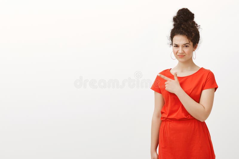 Disgusted european female in stylish casual red dress, folding lips and frowning, looking from under forehead while