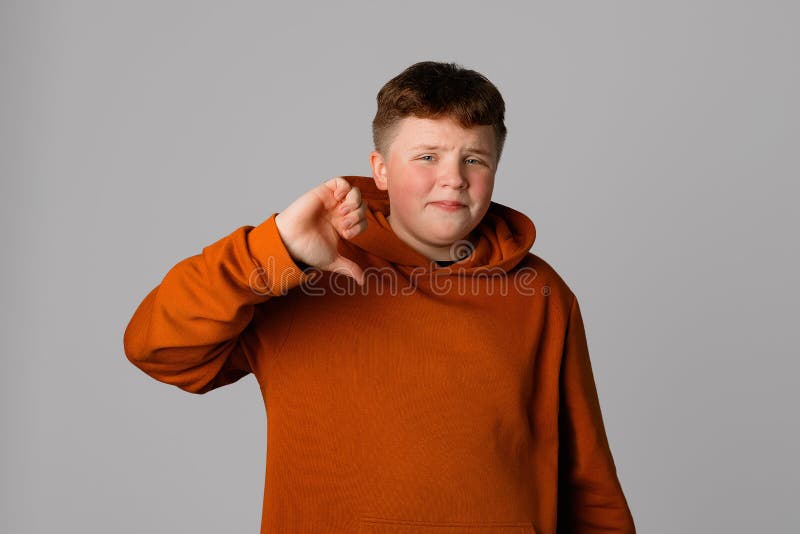 Dissatisfied teenager boy shows thumb down, looking at the camera, standing over gray background