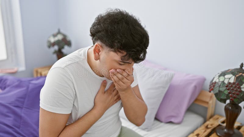 A distressed young man holding his chest and covering his mouth while sitting on a bed in a bedroom stock images