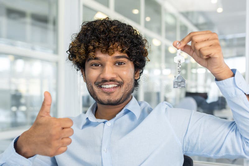 Diverse real estate agent smiling at camera while showing thumb up gesture and holding keys with chain in shape of house. Friendly male recommending buying apartment in new residential complex. Diverse real estate agent smiling at camera while showing thumb up gesture and holding keys with chain in shape of house. Friendly male recommending buying apartment in new residential complex.