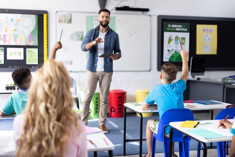 Diverse male teacher and pupils raising hands in elementary school classroom, copy space stock image