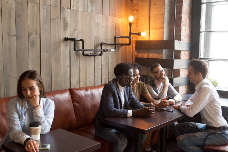 Diverse young friends ignoring sad girl sitting alone in cafe