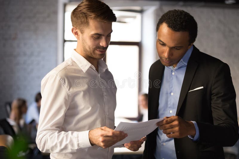 Diverse employees discussing company financial report, statistics, project results, Caucasian businessman holding paper document, supervisor checking work of African American intern in office. Diverse employees discussing company financial report, statistics, project results, Caucasian businessman holding paper document, supervisor checking work of African American intern in office