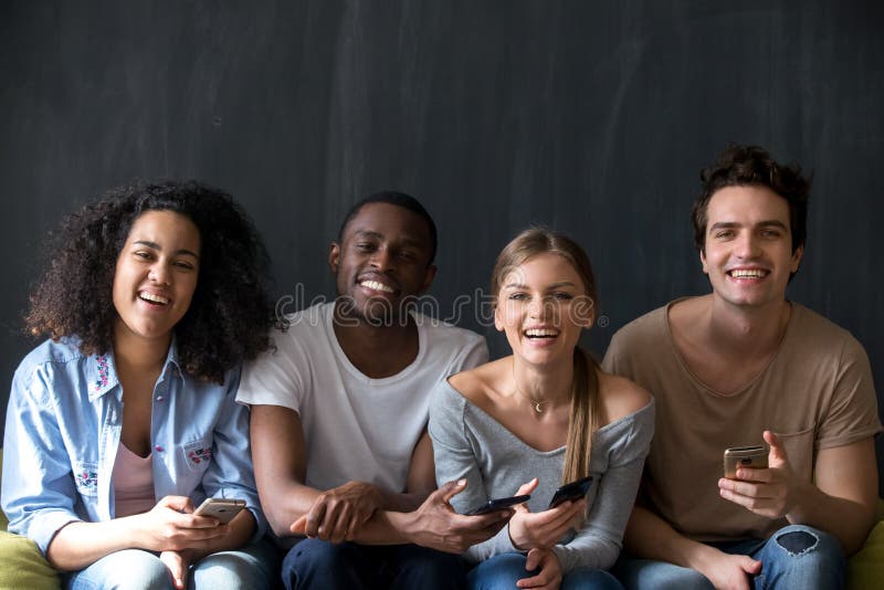 Happy diverse students multiracial friends sitting together indoor. Attractive smiling girls handsome guys holding mobile phones laughing looking at camera. Friendship and wireless technology concept. Happy diverse students multiracial friends sitting together indoor. Attractive smiling girls handsome guys holding mobile phones laughing looking at camera. Friendship and wireless technology concept