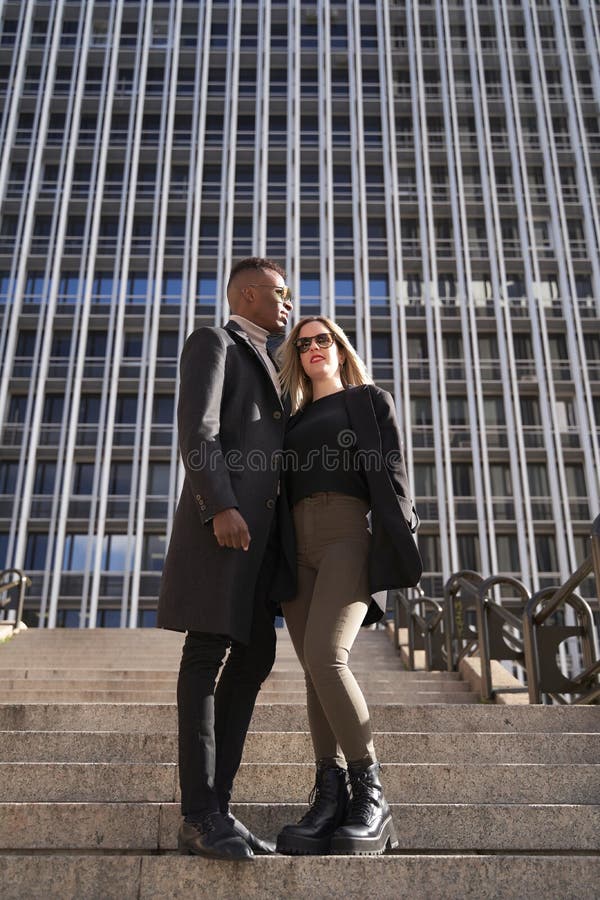 Diversifierat par utanför högrekbyggnaden. Undanstilt multirasistiskt par med elegant ultralusiskt kramper och som står på fotspår utanför modern skyskrapa på soliga dagar i staden