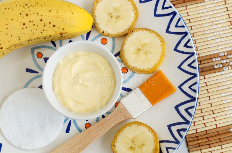 Diy banana mask face cream in the small white bowl, make-up brush, cotton pads and banana slices.