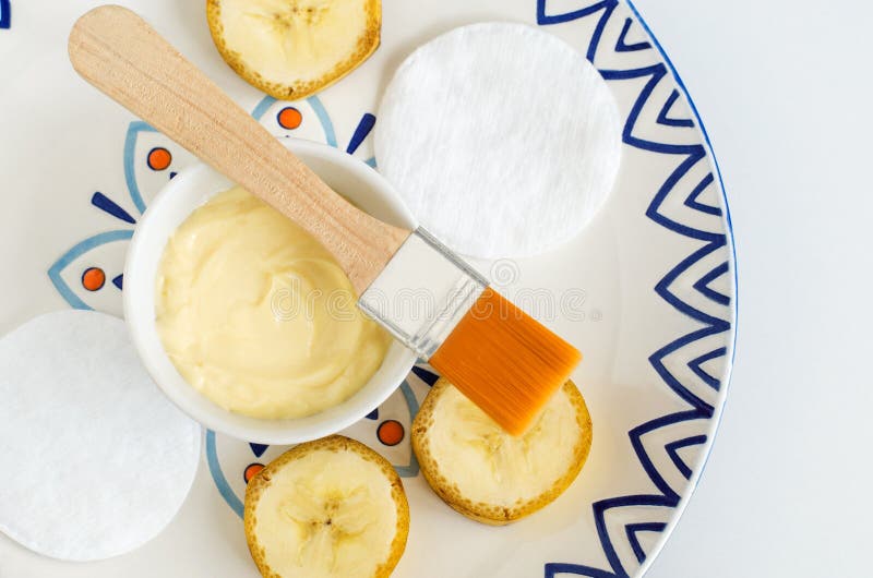 Diy banana mask face cream in the small white bowl, make-up brush, cotton pads and banana slices.