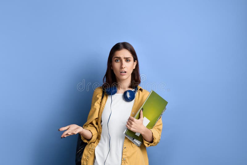 How come, you let me down. Disappointed student girl is throwing hand in indignation, holding books, wearing yellow shirt, white t-shirt, black bag and headphones over neck, on blue background. How come, you let me down. Disappointed student girl is throwing hand in indignation, holding books, wearing yellow shirt, white t-shirt, black bag and headphones over neck, on blue background