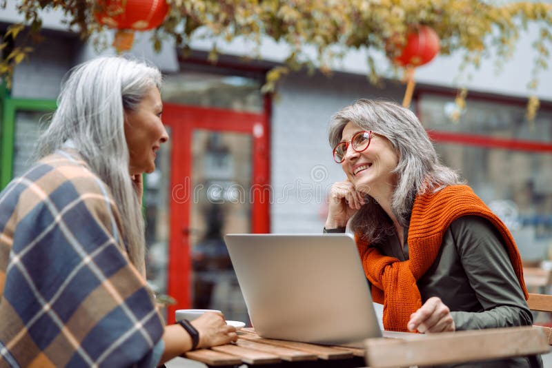 Doamna cu părul gri cu laptop petrece timp cu un prieten asiatic pe terasa cafenelei în aer liber stock photos