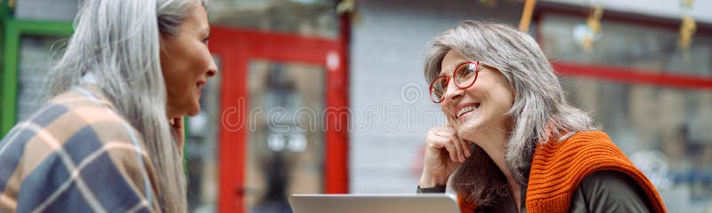 Doamna cu părul gri cu laptop petrece timp cu un prieten asiatic pe terasa cafenelei în aer liber stock photography