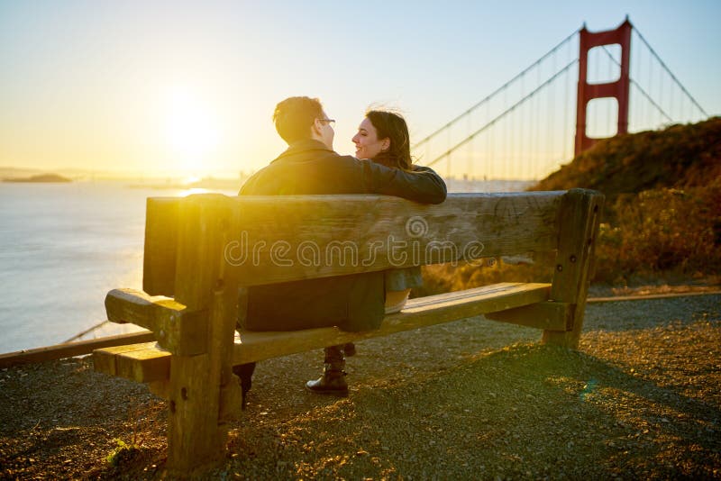 Couple sitting on bench along bay in Golden Gate Park with bridge in background in San Francisco, California at sunset. Couple sitting on bench along bay in Golden Gate Park with bridge in background in San Francisco, California at sunset.