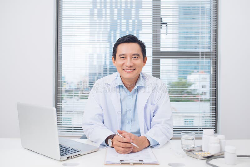 Docteur asiatique de sourire s'asseyant à son bureau dans le bureau médical image libre de droits Docteur asiatique de sourire s'asseyant à son bureau dans le bureau médical image libre de droits