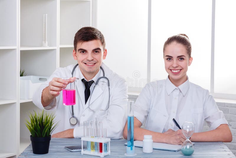 Docteur de deux jeunes dans l'uniforme, se reposant au bureau et souriant, le type tenant un flacon en verre avec un liquide rose photo libre de droits Docteur de deux jeunes dans l'uniforme, se reposant au bureau et souriant, le type tenant un flacon en verre avec un liquide rose photo libre de droits