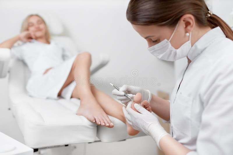 View of female client felaxing in arm chair when podiatrist doctor making procedure for peeling feet with iron tool. Podiatry proffesional service and medical concept. View of female client felaxing in arm chair when podiatrist doctor making procedure for peeling feet with iron tool. Podiatry proffesional service and medical concept.