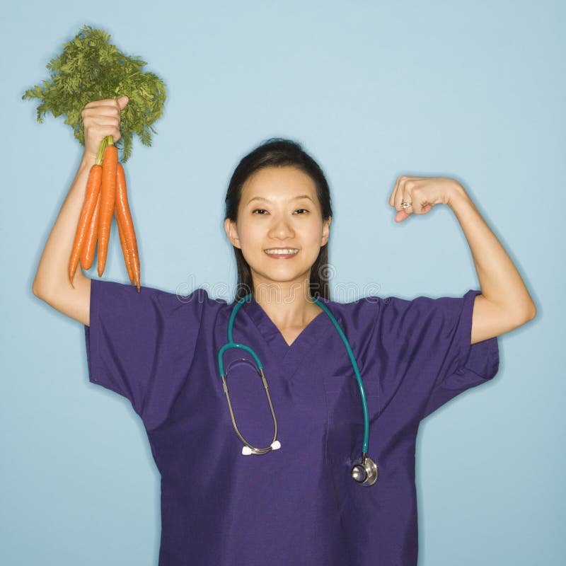 Asian Chinese mid-adult female doctor flexing muscle holding up bunch of carrots against blue background smiling and looking at viewer. Asian Chinese mid-adult female doctor flexing muscle holding up bunch of carrots against blue background smiling and looking at viewer.