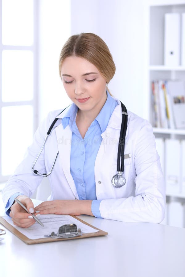 Doctor de la mujer joven en el trabajo mientras que llena encima de expedientes de historia de medicación en oficina del hospital imagenes de archivo Doctor de la mujer joven en el trabajo mientras que llena encima de expedientes de historia de medicación en oficina del hospital imagenes de archivo