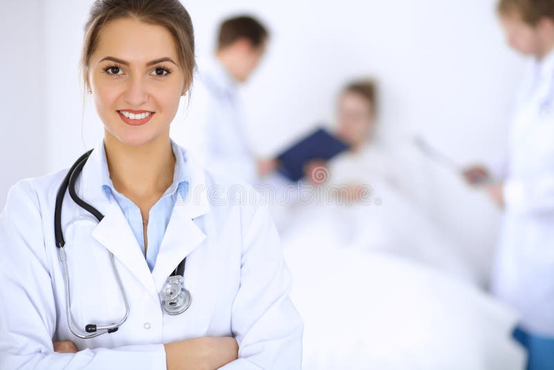 Doctor de sexo femenino que sonríe en el fondo con el paciente en la cama y dos doctores imagen de archivo libre de regalías Doctor de sexo femenino que sonríe en el fondo con el paciente en la cama y dos doctores imagen de archivo libre de regalías