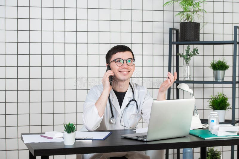 Doctor de sexo masculino que usa el teléfono mientras que trabaja en el ordenador en la tabla en clínica fotografía de archivo libre de regalías Doctor de sexo masculino que usa el teléfono mientras que trabaja en el ordenador en la tabla en clínica fotografía de archivo libre de regalías