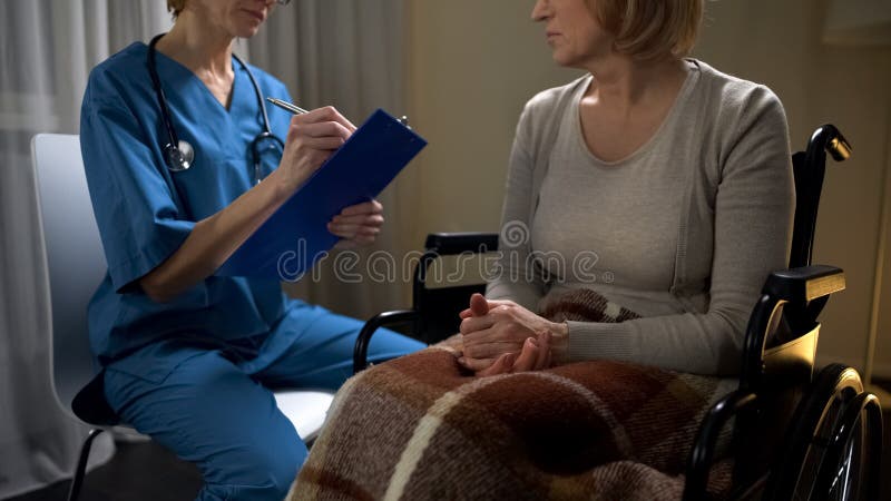 Doctor en llamada que habla con la mujer enferma en silla de ruedas y que hace las notas, diagnosis imagen de archivo Doctor en llamada que habla con la mujer enferma en silla de ruedas y que hace las notas, diagnosis imagen de archivo