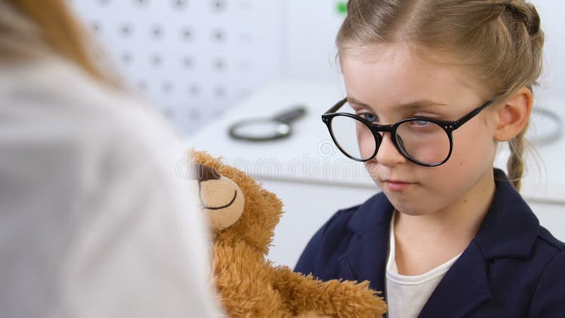 Doctor giving teddy bear to small female patient in glasses, kids clinic service