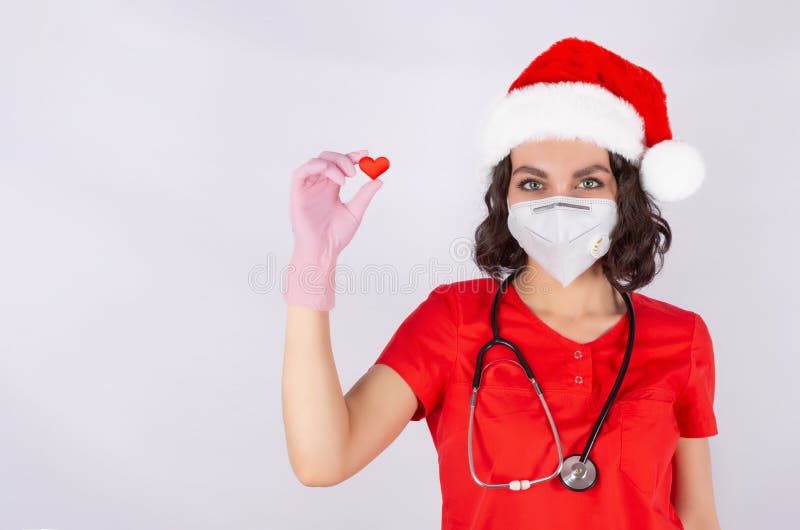 Portrait of a doctor in a medical protective mask, Santa hat and pink nitrile gloves, hand showing a heart, symbol of love, cardiology isolated on a white background. Copy space