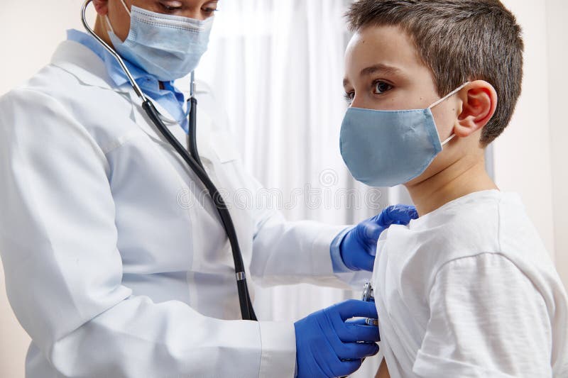 Doctor pediatrician in medical uniform uses a stethoscope while auscultating a little boy in protective medical mask