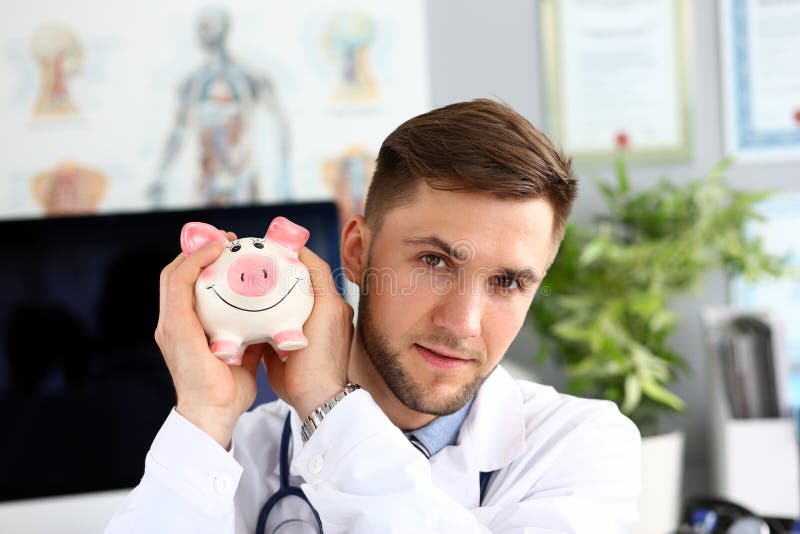 Portrait of smart doc holding and shaking thrift-box. Thoughtful physician sitting in polyclinic cabinet and looking at camera with certainty. Health insurance policy concept. Blurred background. Portrait of smart doc holding and shaking thrift-box. Thoughtful physician sitting in polyclinic cabinet and looking at camera with certainty. Health insurance policy concept. Blurred background