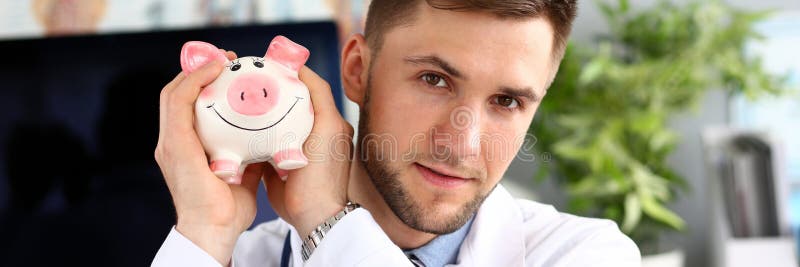 Portrait of smart doc holding and shaking thrift-box. Thoughtful physician sitting in polyclinic cabinet and looking at camera with certainty. Health insurance policy concept. Blurred background. Portrait of smart doc holding and shaking thrift-box. Thoughtful physician sitting in polyclinic cabinet and looking at camera with certainty. Health insurance policy concept. Blurred background