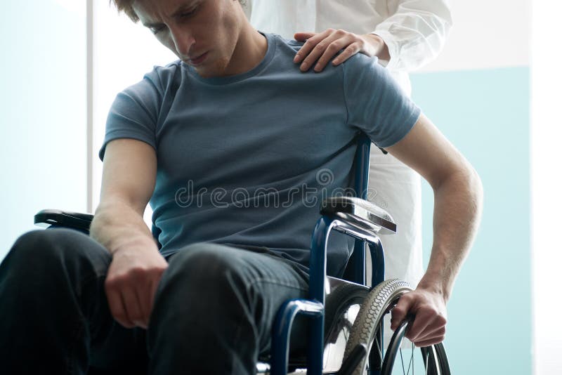 Doctor que consuela al hombre joven que se sienta en silla de ruedas fotos de archivo Doctor que consuela al hombre joven que se sienta en silla de ruedas fotos de archivo