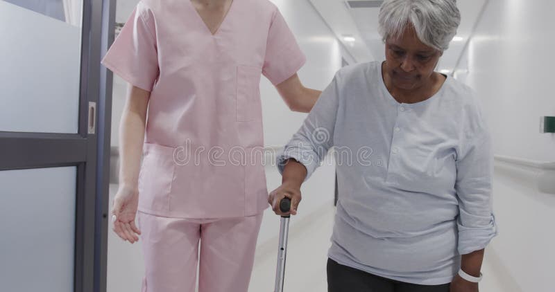 Doctora diversa hablando con paciente de edad avanzada caminando con palo en cámara lenta en el corredor almacen de video