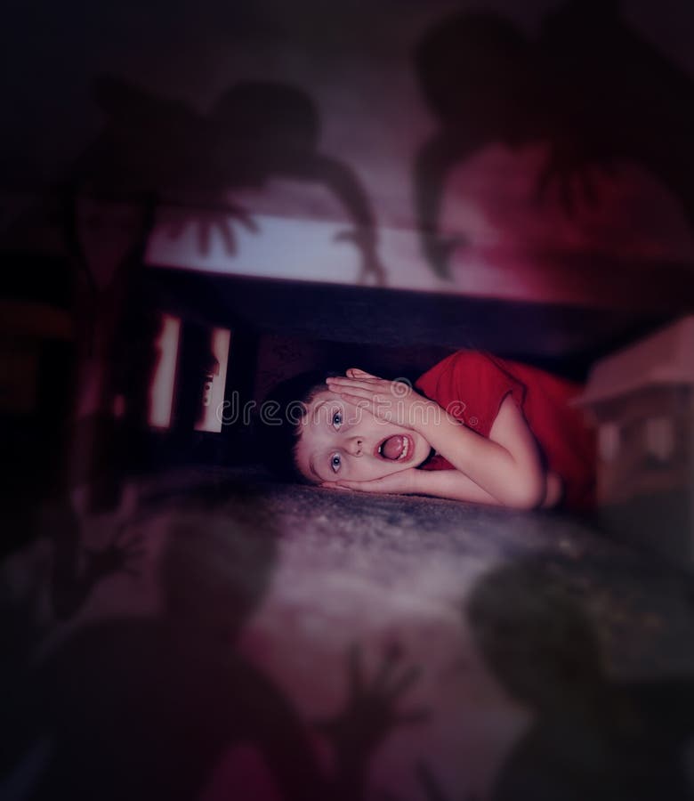 A young scared boy is hiding under a bed looking at black scary monster ghosts in fear for a bedtime or evil concept. A young scared boy is hiding under a bed looking at black scary monster ghosts in fear for a bedtime or evil concept.