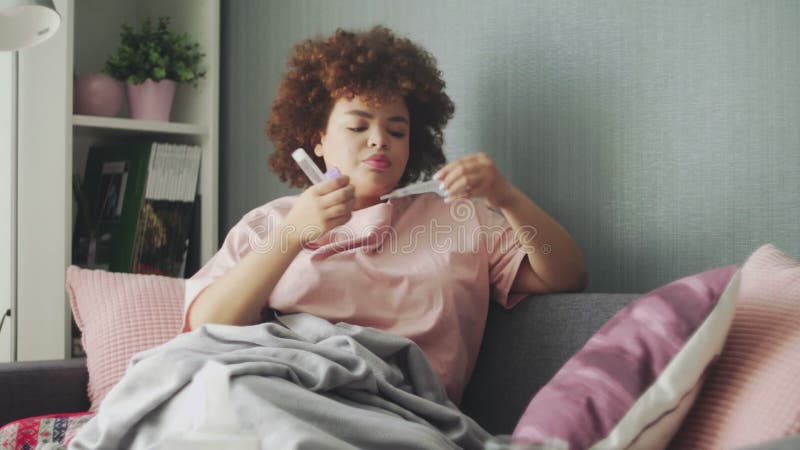 Doente da américa africana, com gripe no sofá em casa. jovem negra com frio verificando a temperatura com