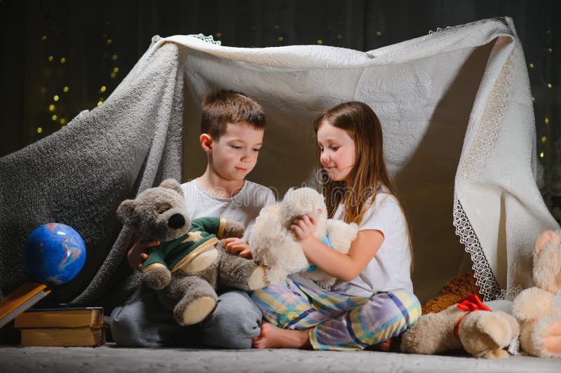 Doi copii mici se joacă acasă seara pentru a construi un cort de camping pentru a citi cărți cu lanternă și a dormi înăuntru. conc stock images