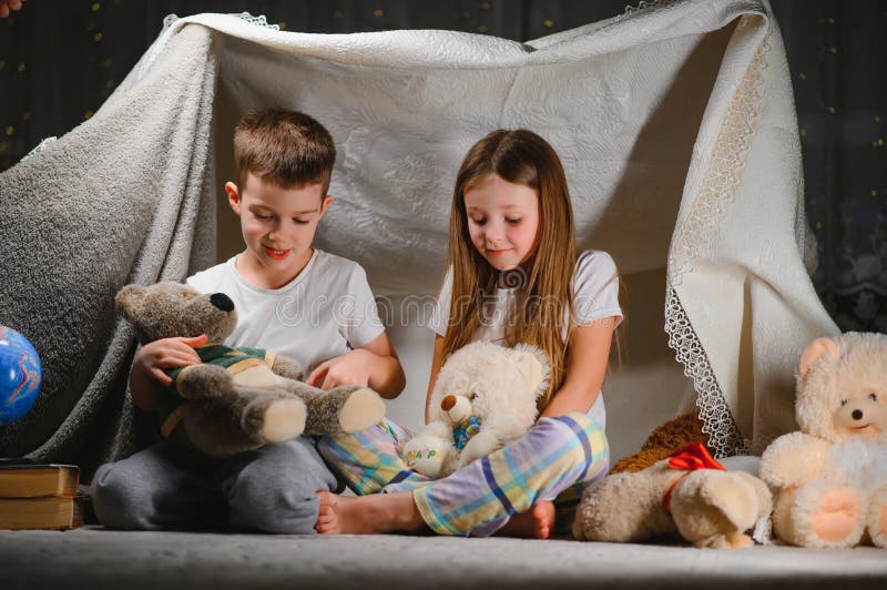Doi copii mici se joacă acasă seara pentru a construi un cort de camping pentru a citi cărți cu lanternă și a dormi înăuntru. conc stock photo